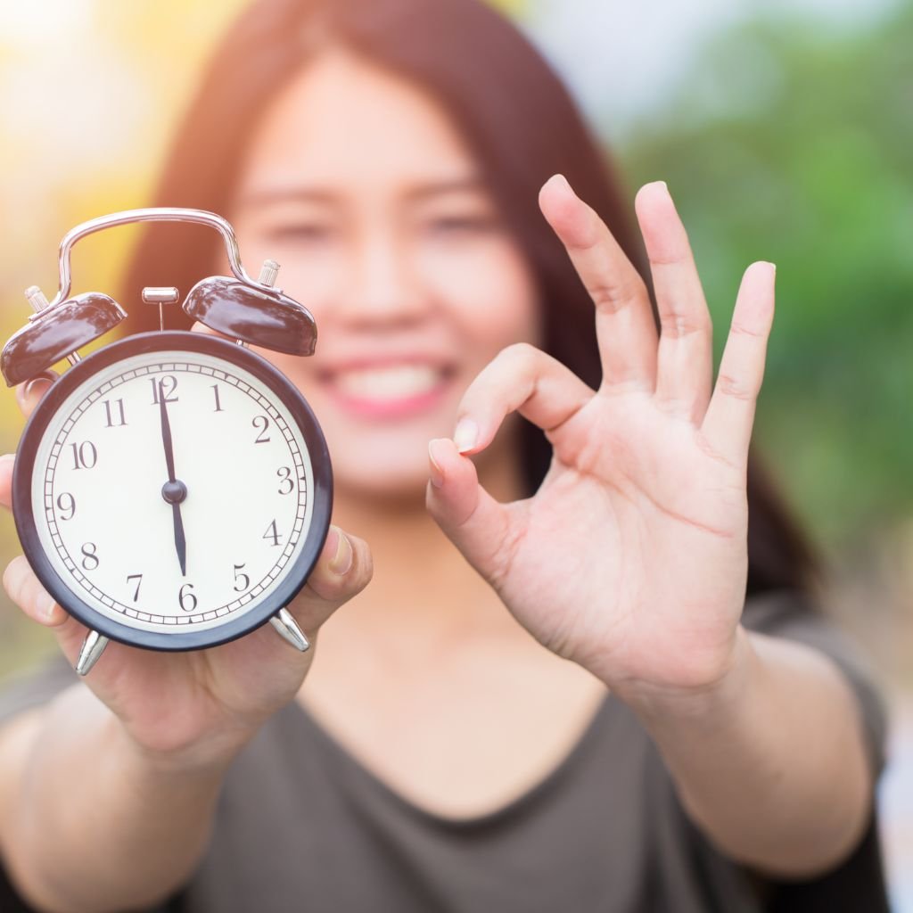 A woman smiling brightly holds up a classic black and white alarm clock showing the time as 7 o'clock. With her other hand, she makes an 'OK' gesture, symbolizing the importance of choosing the right time. The image conveys positivity and the concept of timing.