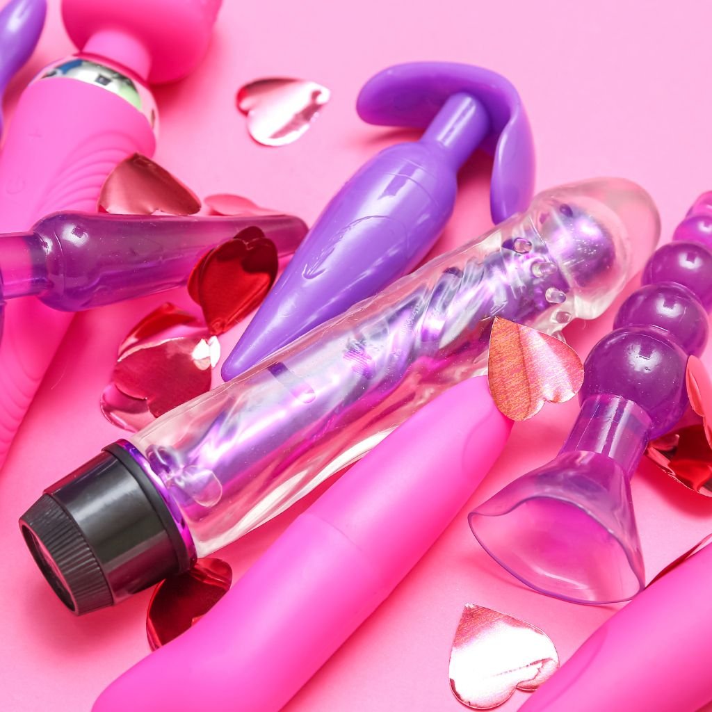 The image shows a selection of brightly coloured sex toys, mostly in shades of pink and purple, arranged on a pink background. The toys include a transparent vibrator, a couple of differently shaped butt plugs, and a few vibrators with varying textures and designs. Scattered around the toys are shiny, red, heart-shaped confetti, adding a playful and romantic touch to the image. The overall feel is fun and vibrant, with an emphasis on variety in shape and texture.