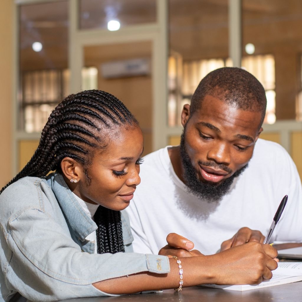 A couple sits closely together, engaged in a shared activity. The man is writing with a pen while explaining something to the woman, who listens attentively. Both are smiling slightly, creating an intimate and collaborative atmosphere, symbolizing exploring or learning together.