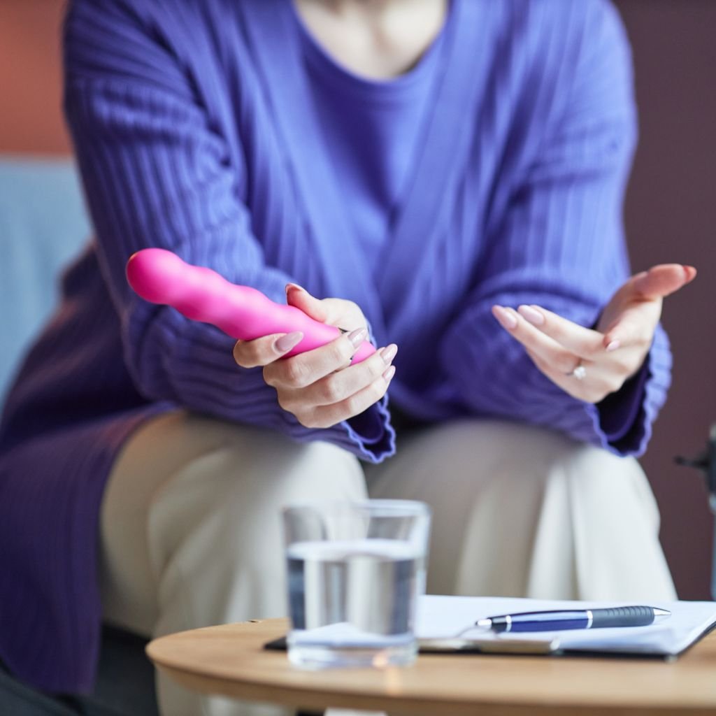 The image appears to show a person sitting while holding a pink, rounded vibrator in their hands. The individual is wearing a blue sweater and is possibly in the middle of a conversation or explaining something, as their hands are expressive. On the table in front of them is a glass of water, a notebook, and a pen, suggesting a casual, perhaps instructional or conversational setting.