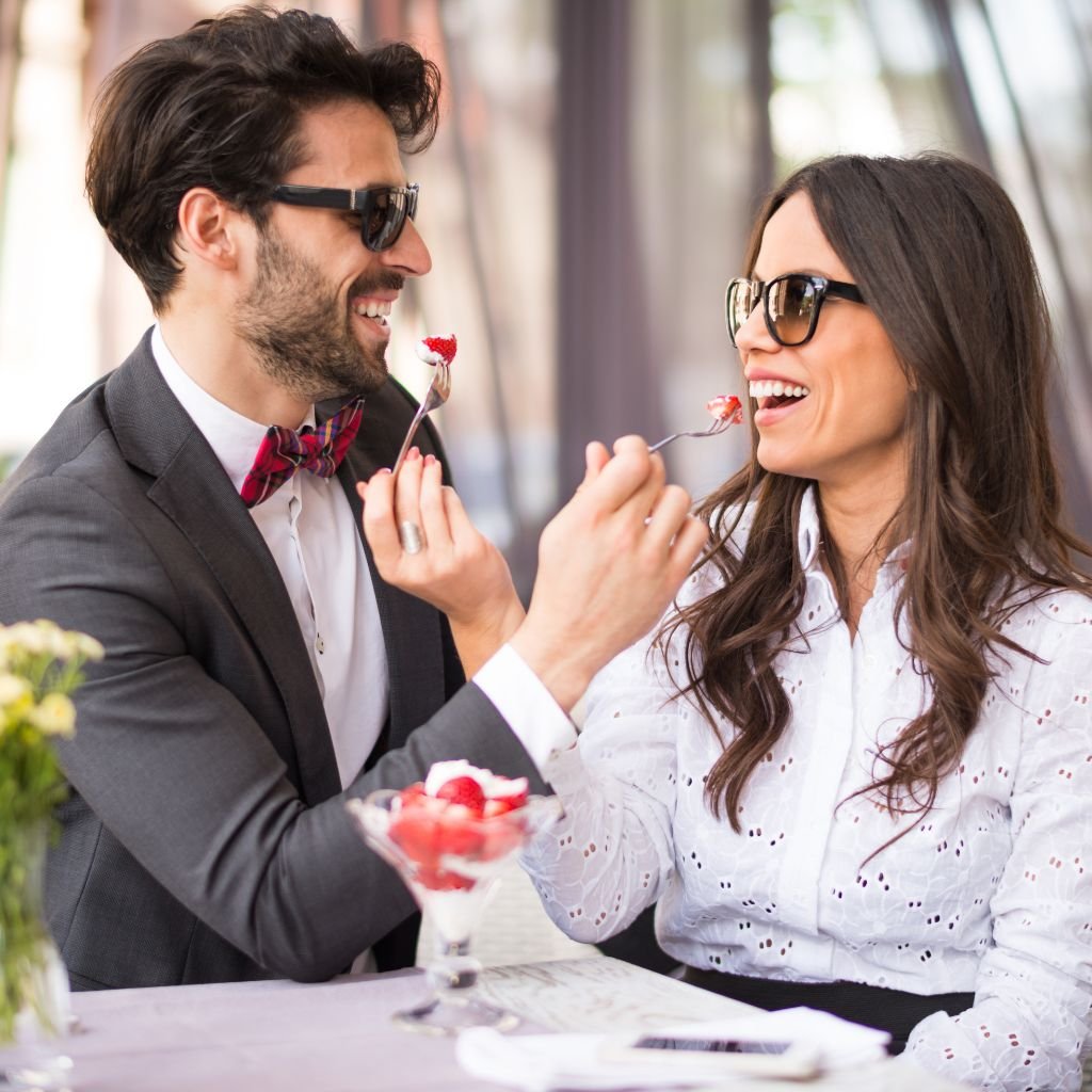 A stylish couple sitting outdoors, sharing dessert while smiling at each other. Both are wearing sunglasses and playfully feeding each other, showing mutual enjoyment and connection. The image represents the concept of mutual pleasure and shared moments in relationships.