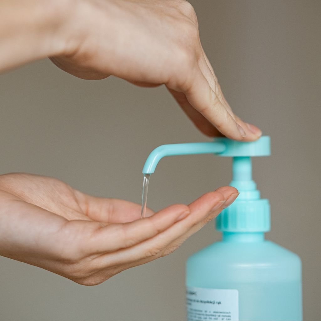 The image depicts a person using a pump dispenser to apply liquid onto their hand, with the liquid being dispensed from a light blue bottle. The focus is on the hands, one pressing the pump and the other receiving the liquid, likely hand sanitizer or soap. This imagery represents cleanliness, safety, and hygiene, highlighting the importance of personal care and health precautions. The neutral background ensures that the attention remains on the hands and the act of dispensing, reinforcing the hygienic theme.