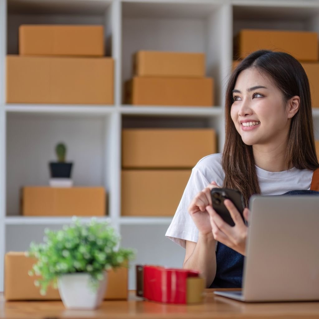 A young woman smiles while holding her phone, seated at a desk with a laptop in front of her. She appears relaxed and content, surrounded by cardboard boxes on shelves behind her, suggesting she is managing a small online business or preparing orders. A small potted plant adds a touch of greenery to the workspace.